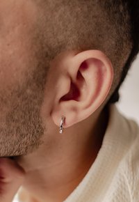 Pendiente de aro plateado en una oreja perforada. El diseño es circular con una superficie lisa y brillante y una ligera hendidura. Cabello corto y bien recortado.