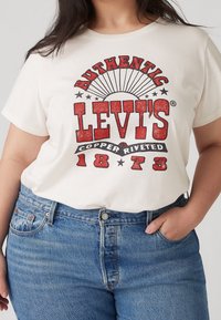 Žena v bílem tričku Levi's s červeným a černým potiskem "Authentic Levi's Copper Riveted 1873" a modrých džínách, ruka v kapse.