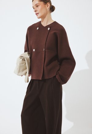 Woman wearing brown buttoned jacket and loose black pants, holding a folded beige bag, against a plain light background.