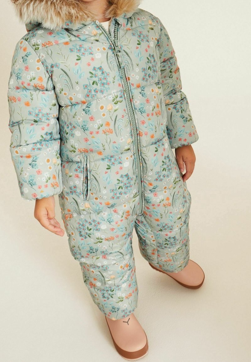 Enfant portant une combinaison de neige matelassée bleu clair à fleurs avec capuche bordée de fourrure et bottes roses, debout sur un fond neutre.