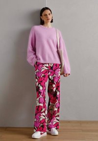 Ljusrosa, oversized stickad tröja i kombination med utsvängda byxor med blommönster i rosa och vitt. Modellen bär vita sneakers och håller en väska.