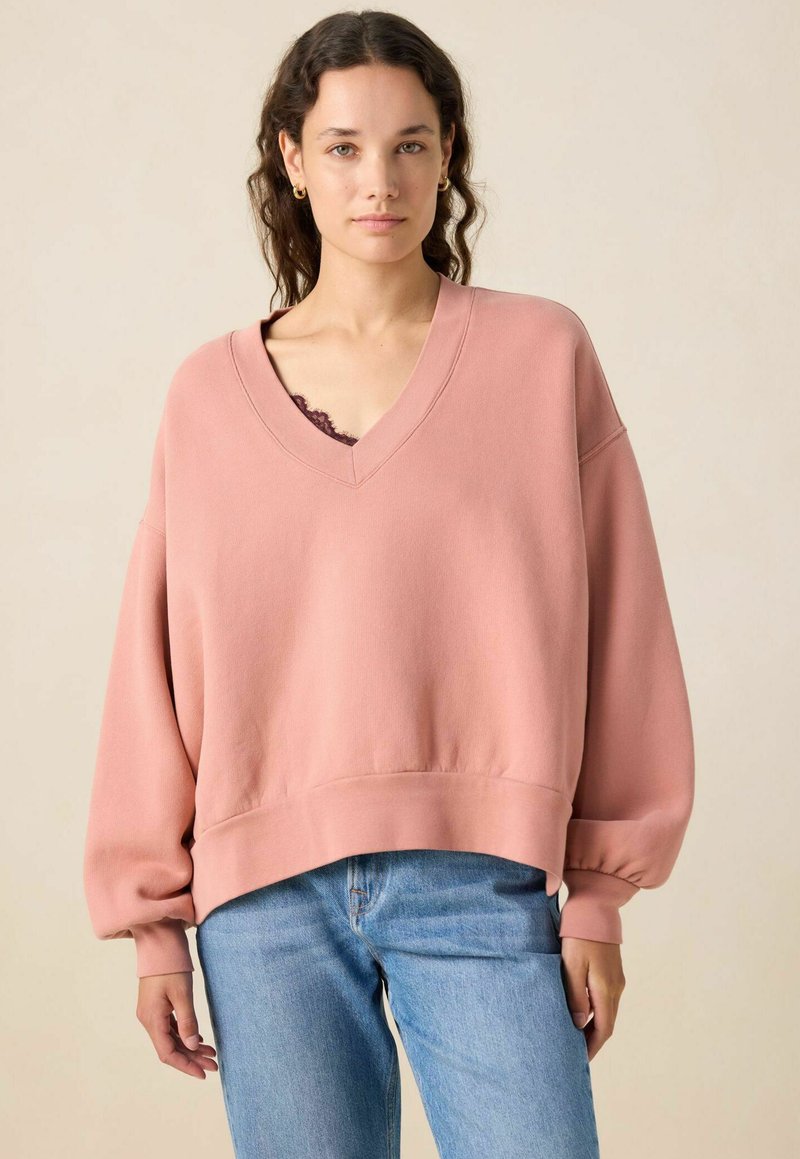 Sweatshirt rose surdimensionné avec un col en V et des manches longues ajustées. Fabriqué en tissu doux, il présente un ourlet et des poignets côtelés, associé à un jean bleu clair.