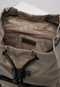 Kellopussin muotoinen beige kangaslaukku, jossa on musta kiristysnaru ja metalliosat. Sisältä löytyy vetoketjullinen tasku ja sileä vuori. Huomaamaton brändäys on näkyvissä.