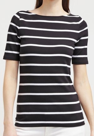 Femme portant une chemise noire à manches courtes avec des rayures blanches horizontales régulièrement espacées et un pantalon blanc.