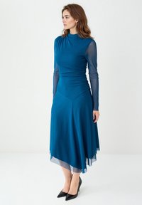Robe teal avec des manches longues et transparentes, un corsage ajusté, et un ourlet évasé plus long à l'arrière. Associée à des escarpins noirs.