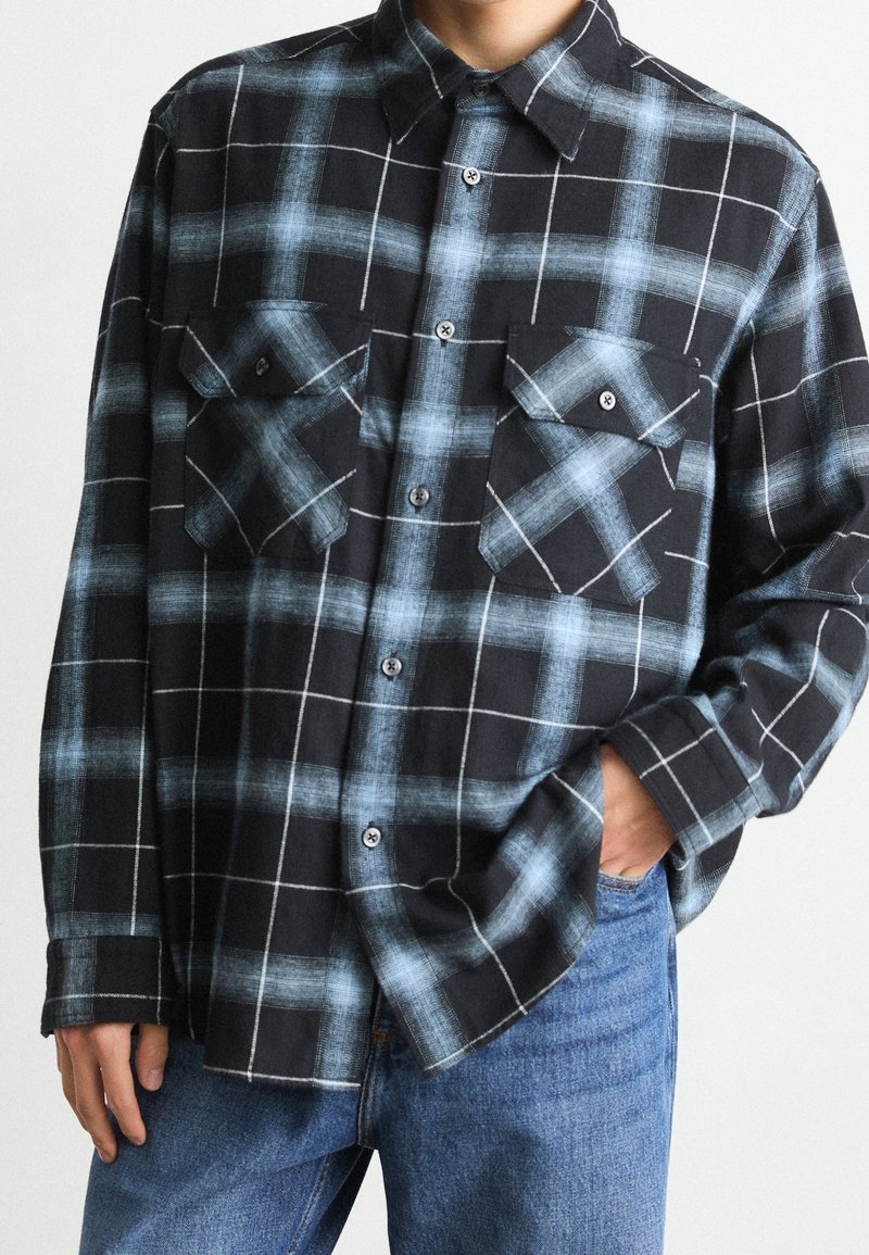Chemise à carreaux noire et bleue à boutons, avec deux poches avant boutonnées, portée non rentrée sur un jean bleu, une main dans la poche.