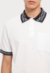 Biely polo tričko s plochým čiernym golierom s reťazovým vzorom a logom. Obsahuje náprsné vrecko a zapínanie na dva gombíky.