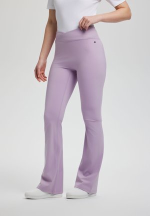 Kobieta w lawendowych spodniami z szerokimi nogawkami z wysokim stanem i białych wsuwanych butach, poprawiająca pasek, na jasnym, jednolitym tle.