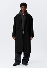 Joven con un abrigo largo negro sobre una sudadera con capucha oscura, jeans oscuros y sueltos, y zapatos negros con textura, de pie frente a un fondo blanco liso.