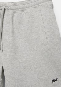 Lysegrå joggingbukser med elastisk talje, snor, sidelomme og lille sort broderet logo nær kanten.