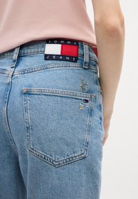Jean en denim bleu clair avec broderies florales sur les poches arrière et une ceinture de marque Tommy Jeans dans des couleurs rouge, blanc et bleu marine.