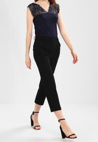 Námořnický top s krajkovým lemem, doplněný černými kalhotami na míru. Modelka má na sobě černé sandály na kotníku. Elegantní, moderní design s přiléhavým siluetou.