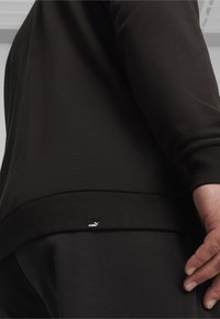 Sweat-shirt noir avec poignets et ourlet côtelés, arborant une petite étiquette logo Puma, présenté de profil, avec un gros plan sur le tissu texturé.