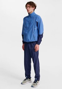 Blå träningsdräkt med en zip-up jacka med marinblå ärmar, ljusblå accenter och matchande byxor med vit kantning. Texturerat tyg, atletisk passform.