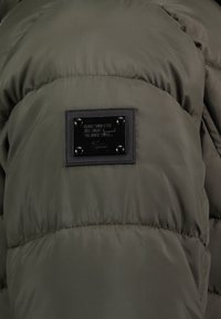 Olijfgroene gewatteerde jas met een klein zwart rechthoekig genaaid etiket met reliëftekst en vier klinknagels.