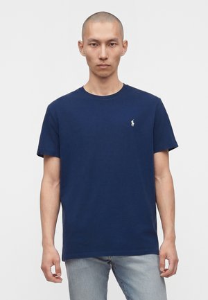 Homme à la tête rasée portant un polo bleu marine à manches courtes avec un logo blanc sur la poitrine et un jean bleu clair, face à l'avant.