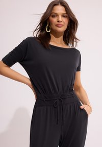 Czarny kombinezon z krótkimi rękawami, elastycznym pasem, detalami ze sznurkiem i bocznymi kieszeniami; gładka tekstura i luźny krój.