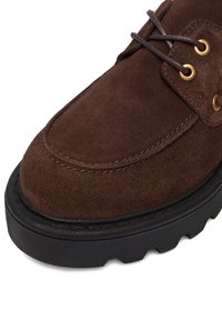 Brūna zamšādas apava ar apaļu purngalu, zelta acuļiem un brūnām auklām. Tam ir teksturēta melna gumijas zole ar biezas protektora dizainu.