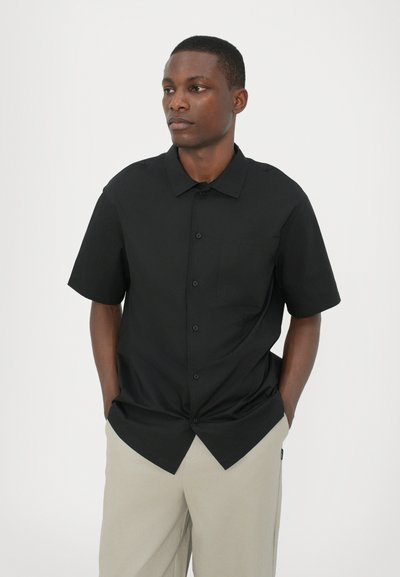 Black short-sleeve shirt with a button-up design, single chest pocket, and smooth texture, paired with light beige trousers.