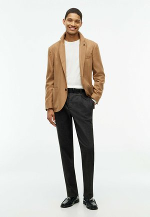 Ung mand smilende iført en beige blazer, hvid skjorte, mørkegrå bukser og sorte pæne sko, står med den ene hånd i lommen.