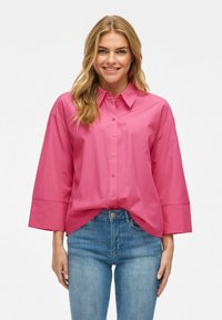 Camicia rosa oversize con bottoni frontali, colletto e maniche larghe. Tessuto liscio con una consistenza fresca, abbinata a jeans di denim blu.