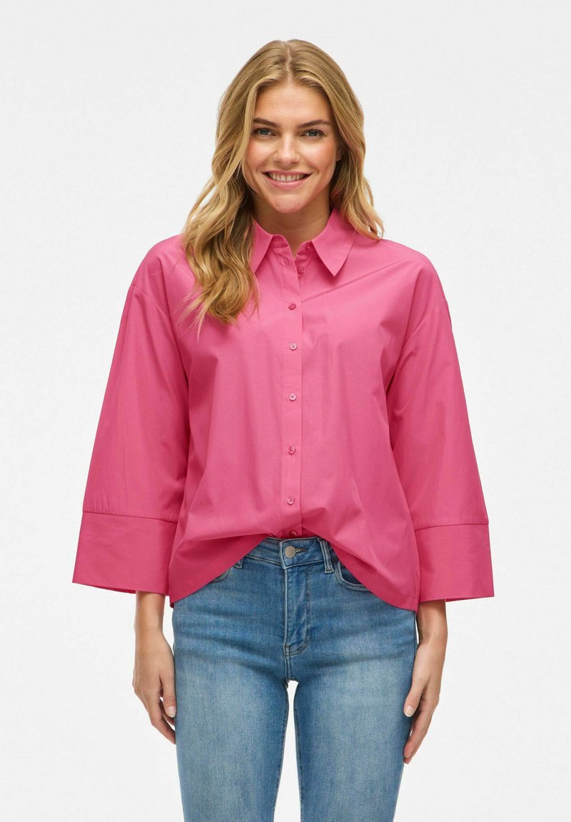 Camicia rosa oversize con bottoni frontali, colletto e maniche larghe. Tessuto liscio con una consistenza fresca, abbinata a jeans di denim blu.