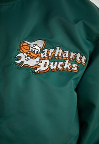 Grøn jakke med et fremtrædende broderet logo med en and og en basketball, accentueret med orange og hvide detaljer. Glat stof tekstur.