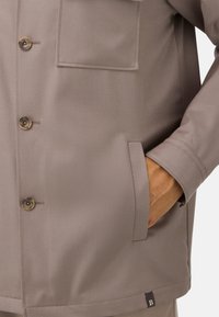 Grå knäppt jacka i slät tyg, med en bröstficka, sidofickor och en logotyp på fållen.