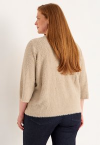 Helle beige, flauschige Strickjacke mit Dreiviertelärmeln, kombiniert mit dunkelblauen Jeans. Zeigt eine weiche Textur und eine lockere Passform.
