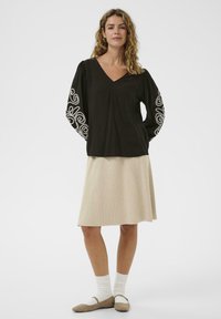 Femme aux cheveux bouclés portant un blouse noire avec une broderie blanche sur les manches, une jupe beige jusqu'aux genoux, des chaussettes blanches et des ballerines beiges.