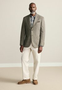 Sacou maro cu model houndstooth, cămașă deschisă cu carouri, pantaloni crem și mocasini maro. Are buton simplu și accent de batistă de buzunar.