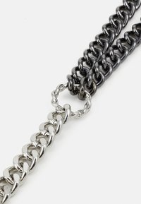 Collier en chaîne métallique avec des maillons noirs et argentés alternés, agrémenté d'un détail de bague de connexion torsadée. Texture lisse dans l'ensemble.