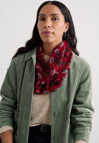 Jauna sieviete, kas valkā sarkanu ziedotu šali, zaļu korduroja jaku, bēša džemperi un nelielus zeltainus apli auskarus uz vienkārša baltā fona.