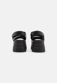 Black platform sandals with adjustable straps, smooth synthetic material, and a textured outsole. Logo embossed on the heel.