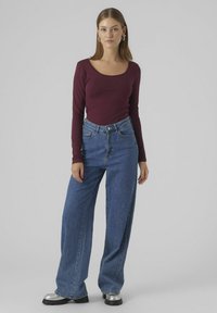 Haut à manches longues bourgogne associé à un jean bleu à taille haute et jambes larges, avec des chaussures à platform argent métallisé, le tout sur un fond neutre.