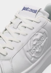 Beli usnjeni superg s belimi vezalkami, reliefnim logotipom "Just Cavalli" na strani in napisom "Just Cavalli" na jezičku v temno modri barvi.