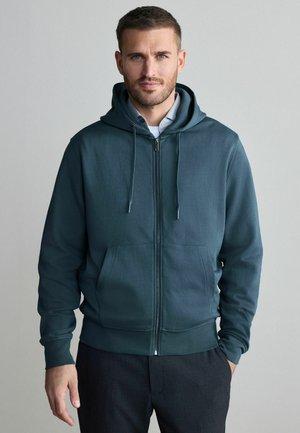 Sudadera con cremallera de color verde azulado oscuro en un tejido suave y liso, con un bolsillo frontal y cordones en la capucha. El modelo la lleva sobre una camisa con cuello.