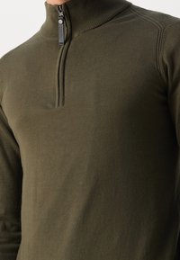 Pull-over à fermeture éclair couleur vert olive en tissu tricoté doux, doté d'un col montant et d'une courte fermeture éclair. Texture lisse avec des détails de coutures minimales.