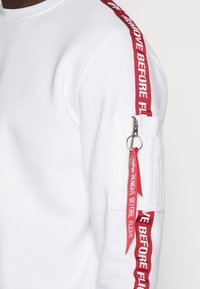 Felpa bianca con accenti rossi, caratterizzata da strisce laterali e una tasca con zip con un'etichetta rossa "RIMUOVERE PRIMA DEL VOLO". Texture morbida.