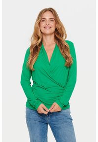 Donna sorridente con lunghi capelli biondi indossa un top avvolgente a maniche lunghe verde e jeans blu, in piedi contro uno sfondo bianco uniforme.