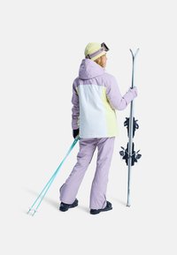 Ubranie narciarskie w kolorze lawendowym i żółtym z białą wstawką, wyposażone w kaptur i rękawice. Osoba trzyma niebieskie kijki narciarskie z przymocowanymi nartami.