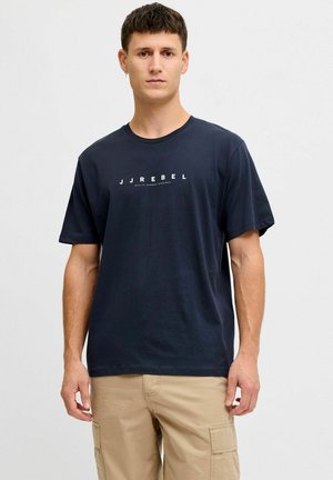 Joven de pie, vistiendo una camiseta azul marino JJREBEL y pantalones cargo beige, fondo neutro.