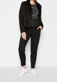 Giacca nera in suede con colletto in pelliccia, t-shirt grigia con grafica, pantaloni neri a sigaretta e sneakers bianche. Borsa nera compatta indossata in vita.