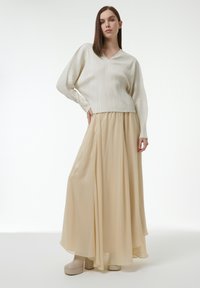 Pull en rib Cord en beige clair associé à une jupe fluide et transparente de couleur beige. Le mannequin porte des chaussures à plateforme chunky beige. Textures lisses, design élégant.