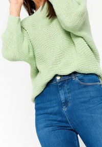 Pull en maille vert clair avec un motif de vagues texturé, associé à un jean bleu ajusté. Le pull a un décolleté large et des épaules tombantes.