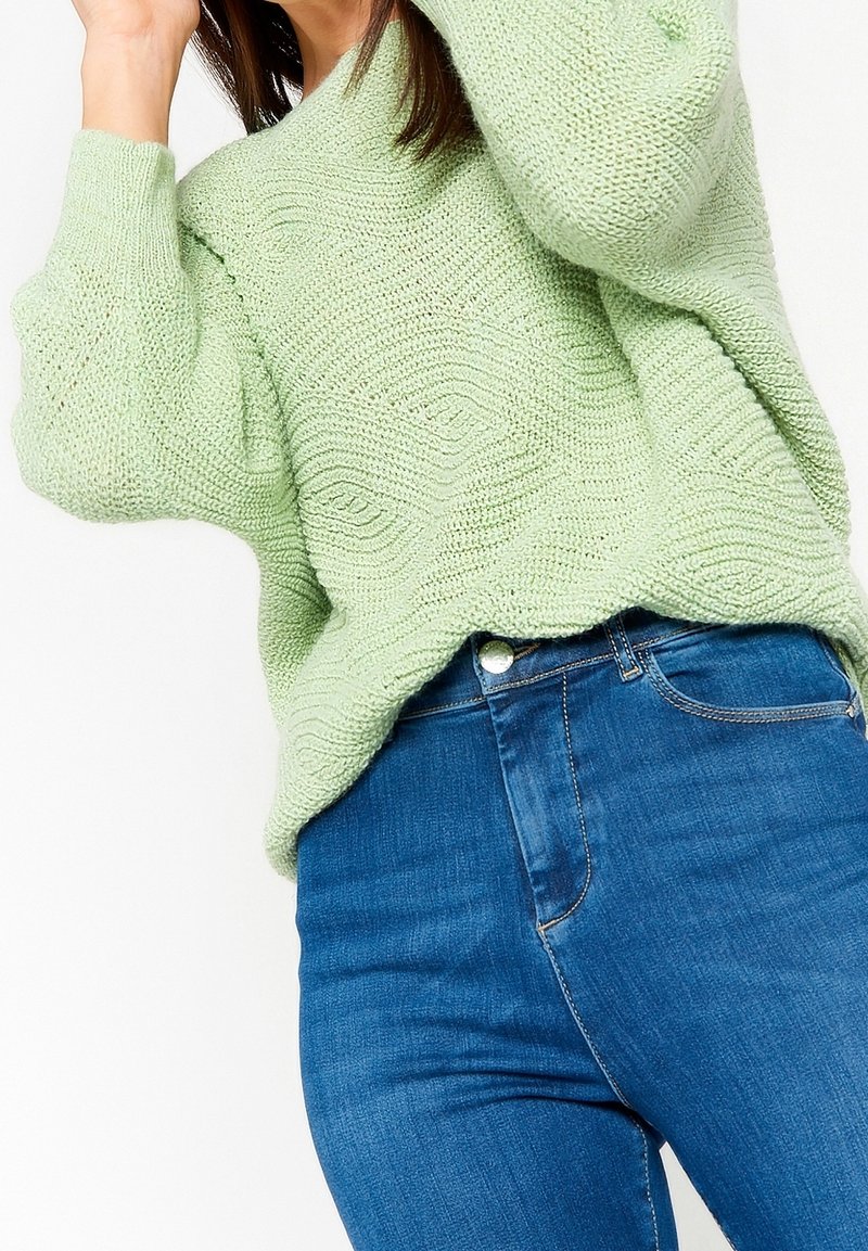 Pull en maille vert clair avec un motif de vagues texturé, associé à un jean bleu ajusté. Le pull a un décolleté large et des épaules tombantes.
