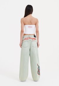 Witte strapless top met rood logo en lichtgroene wijde jeans met een geborduurd ontwerp in rood en blauw op de linkerdij.