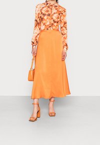 Lange, orange Satinröcke, kombiniert mit einem gemusterten, orange-braunen Hemd mit Knöpfen. Das Outfit umfasst orangefarbene Blockabsatzsandalen und eine kleine Tasche.