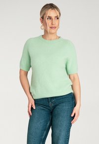 Jasnozielony sweter z krótkim rękawem wykonany z miękkiego materiału; żebrowany dekolt i dół; zestawiony z niebieskimi dżinsami, z naciskiem na luźny fason.