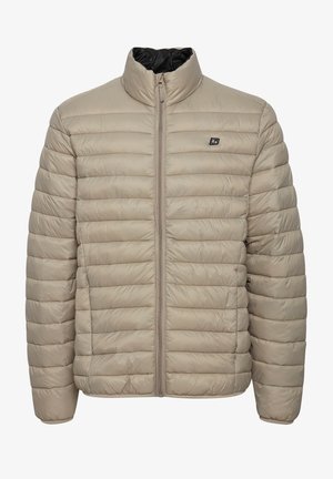 Doudoune beige avec un col montant, une fermeture éclair avant et une texture matelassée en relief. Elle présente un petit logo sur le côté gauche de la poitrine.